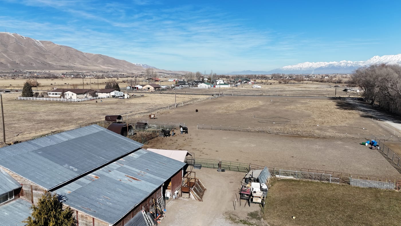 Drone aerial of rural ranch/farm property with barns, animal pens, horses, and mountain backdrop — no solar panels visible; appears to be a pre-installation site survey photo