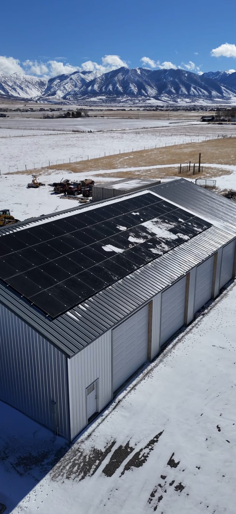 [Tier 1 Trina panels] Stunning vertical drone aerial of metal barn with large rooftop solar array, dramatic snow-covered mountain backdrop, and rural landscape