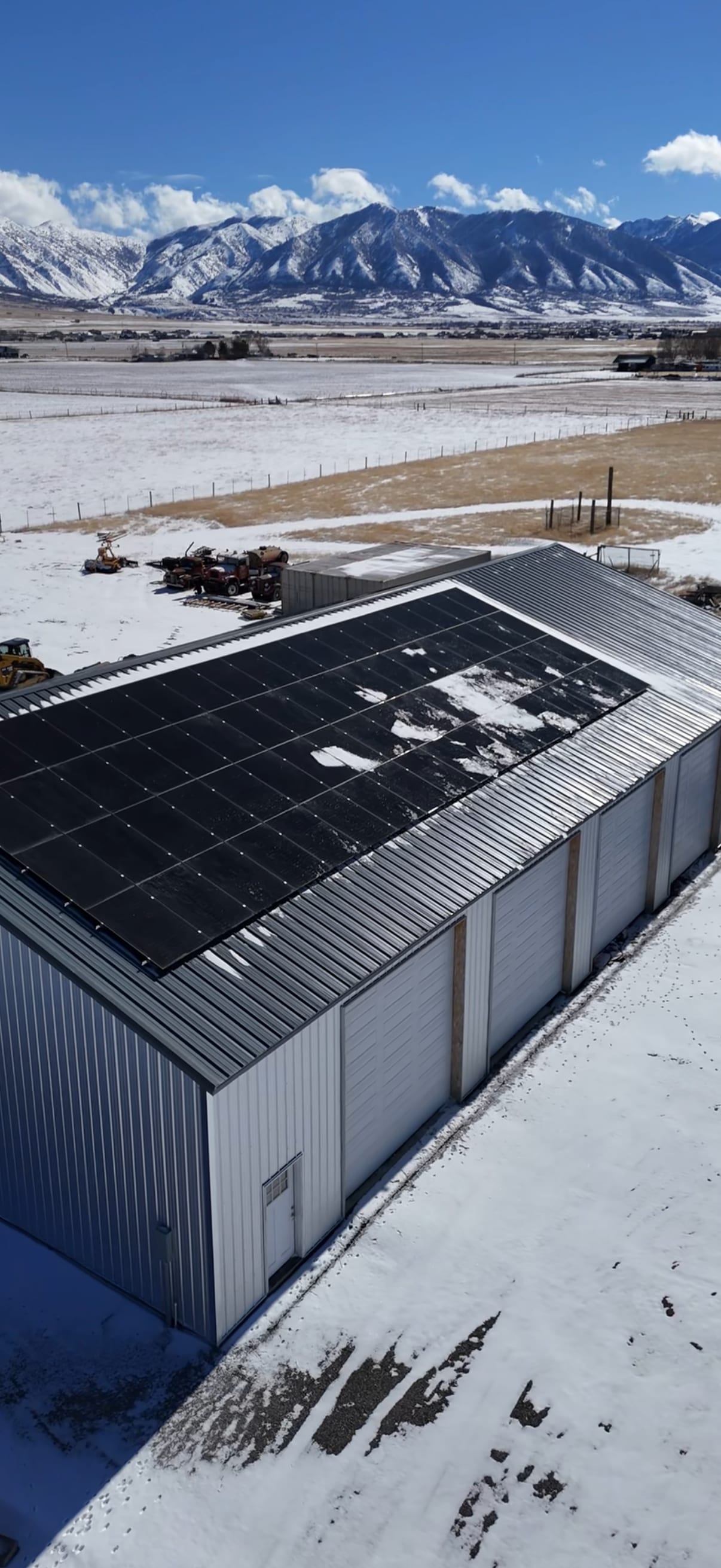 [Tier 1 Trina panels] Stunning vertical drone aerial of metal barn with large rooftop solar array, dramatic snow-covered mountain backdrop, and rural landscape