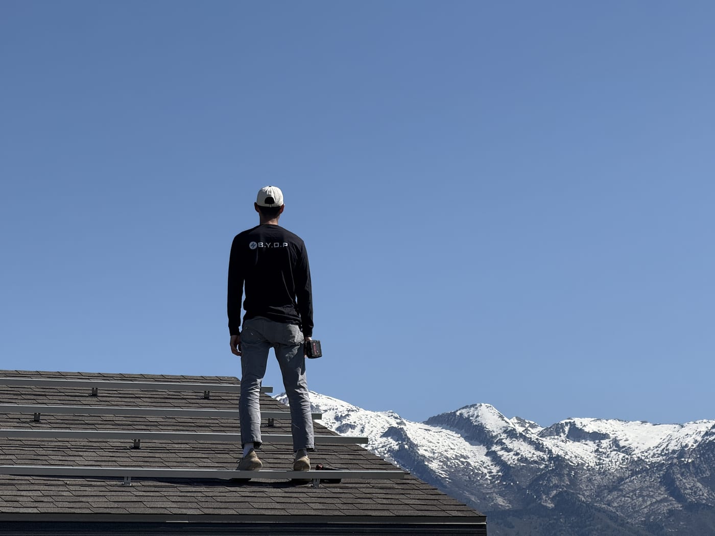 BYOP installer standing on roof next to solar panels with dramatic snow-capped mountains behind