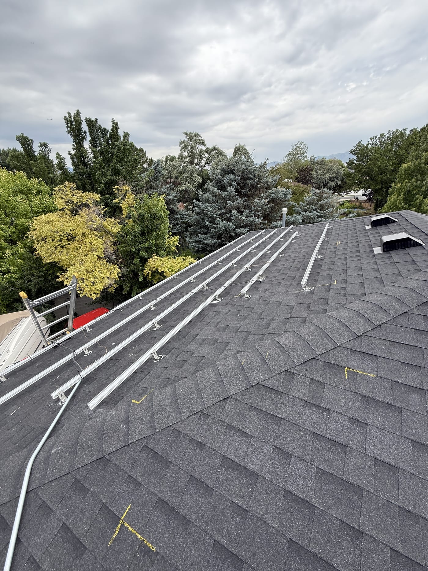 Racking rails installed along shingle roof with ladder and trees in background on cloudy day