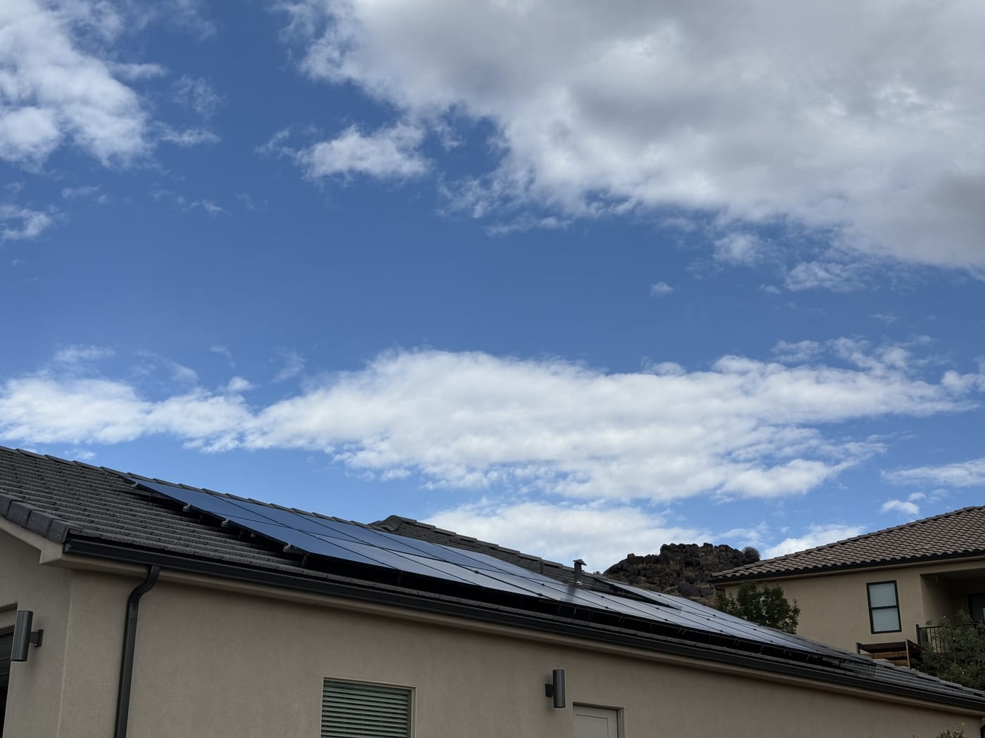 House exterior from ground showing solar panels on roof edge with blue sky and clouds in desert setting