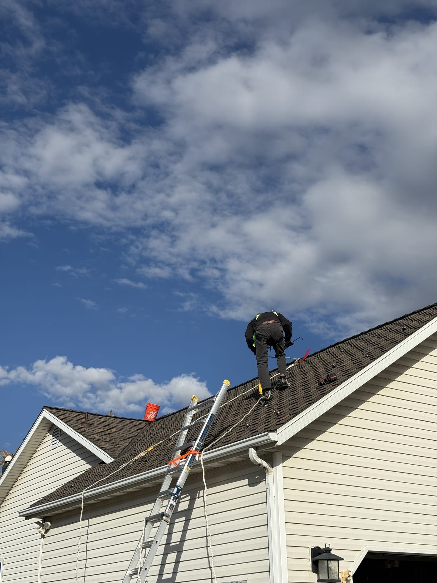 Worker with safety harness and drill on steep shingle roof installing racking mounts against blue sky