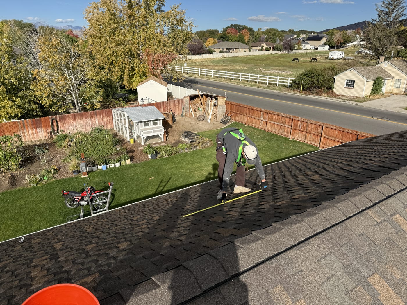 Worker with safety harness on roof edge looking down at backyard with shed and green grass