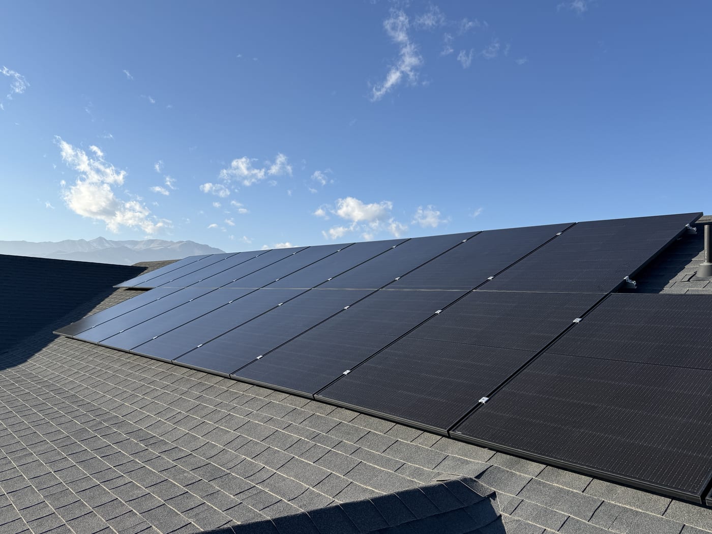 Solar panel array on dark shingle roof from above with blue sky and clouds