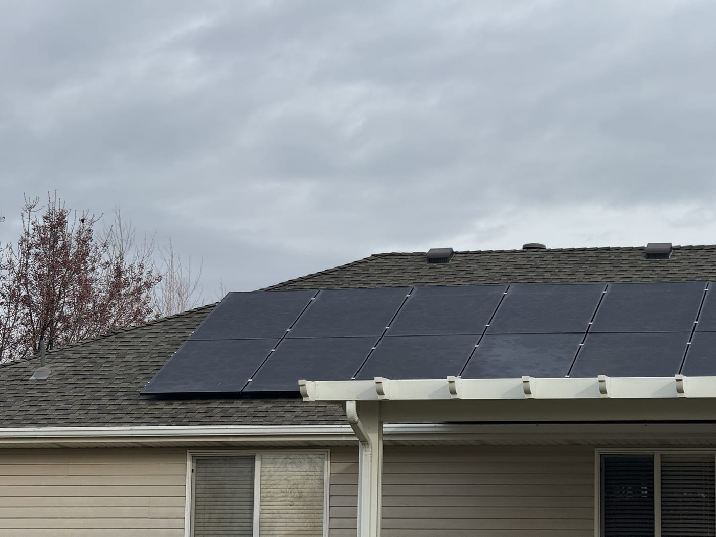 [black-on-black panels] Close-up of black solar panels on gray shingle roof showing racking and clamps, partial house siding visible (image rotated)