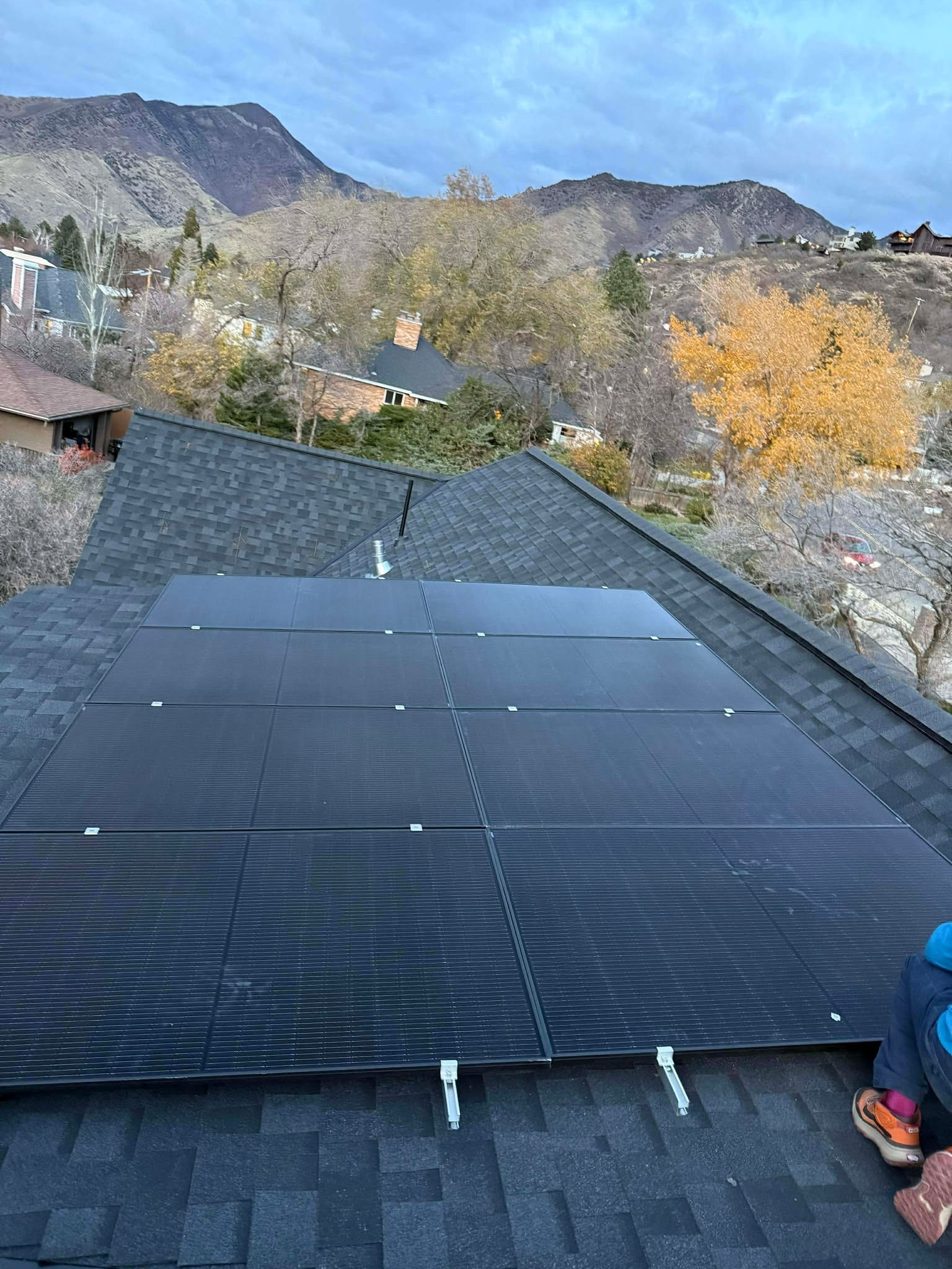 Black solar panels on gray shingle roof with fall foliage and Wasatch mountains behind, shot from roof level