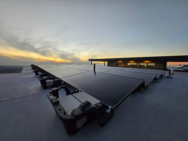 Ballasted solar array on flat roof at sunset, black panels with weighted mounts, valley and mountains in background with golden sky