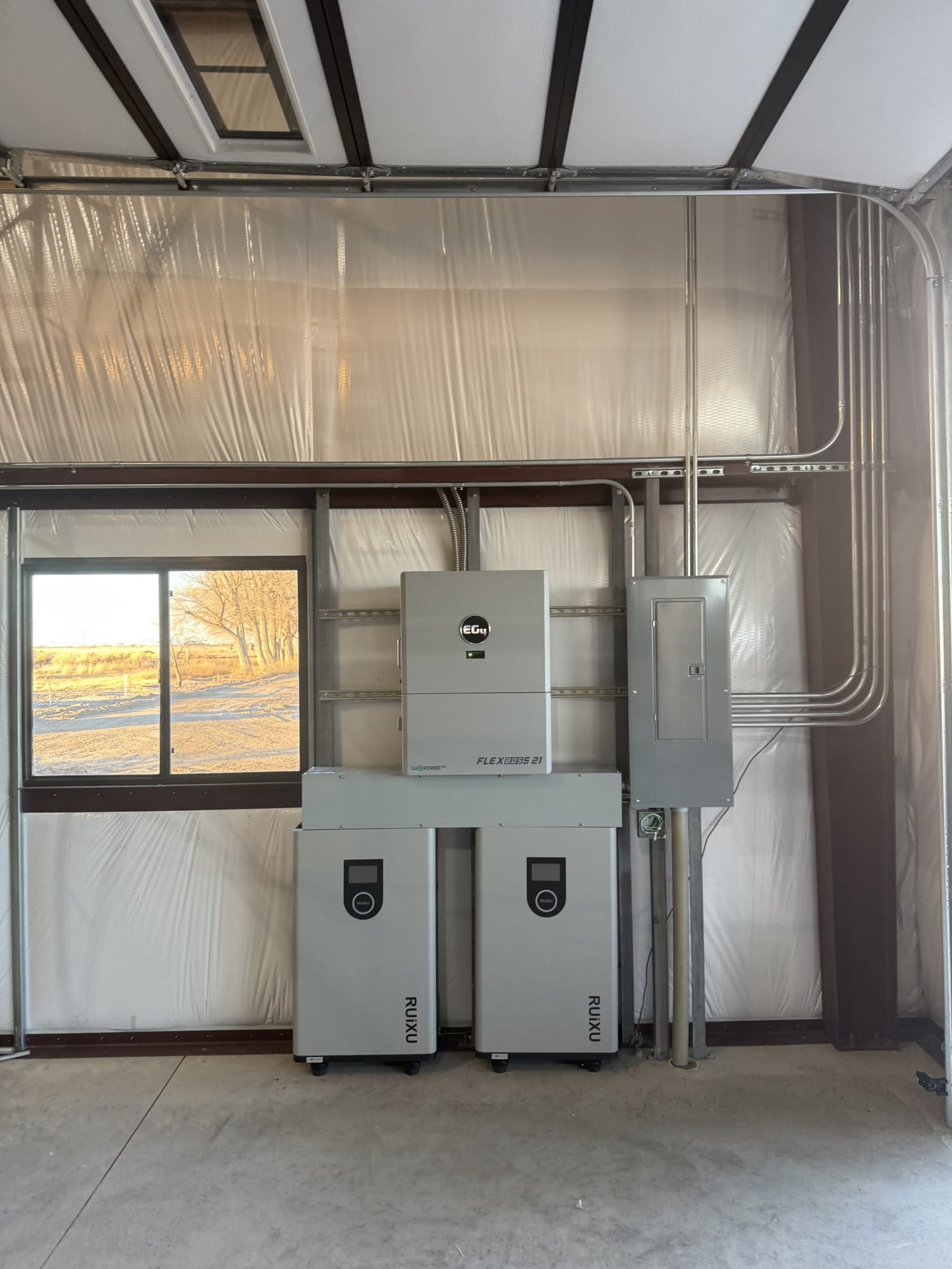 Wide view of EG4 FlexBOSS 21 inverter with two RUiXU batteries, electrical panel, and conduit on garage wall with vapor barrier
