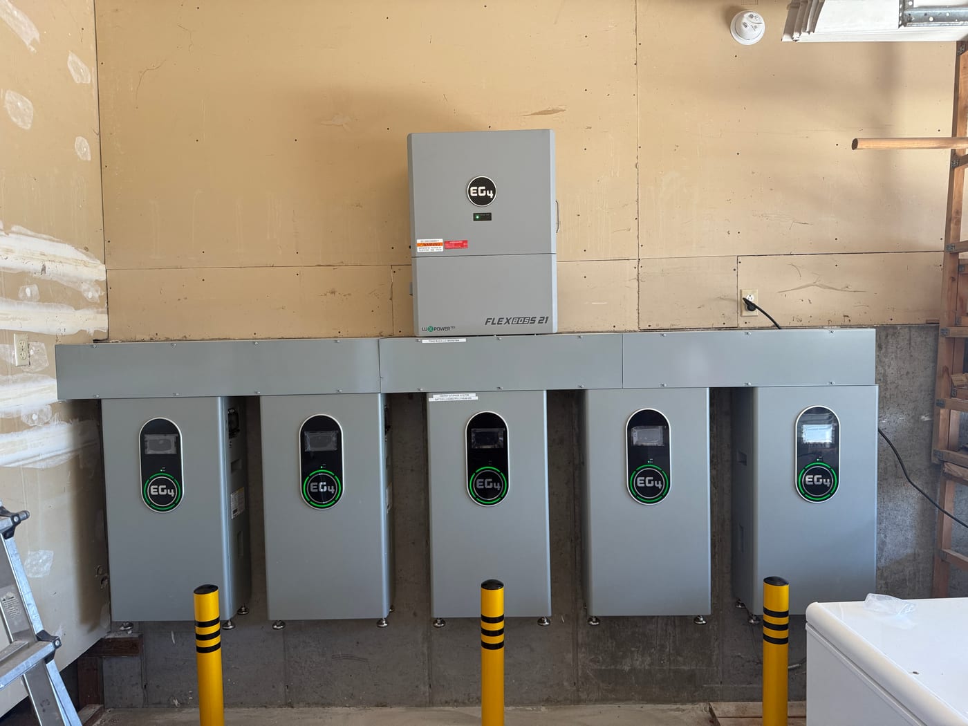 [EG4 FlexBOSS 21] Five EG4 batteries fully mounted on plywood backboard with FlexBOSS 21 inverter below, all green status LEDs lit, yellow conduit guards on top — completed wall (image rotated)