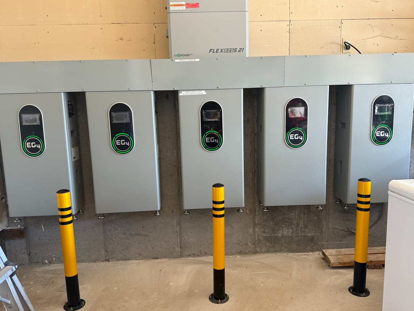 [EG4 FlexBOSS 21] Five EG4 batteries suspended from yellow ceiling-mounted lift columns during installation, FlexBOSS 21 inverter visible below — process shot showing the install method