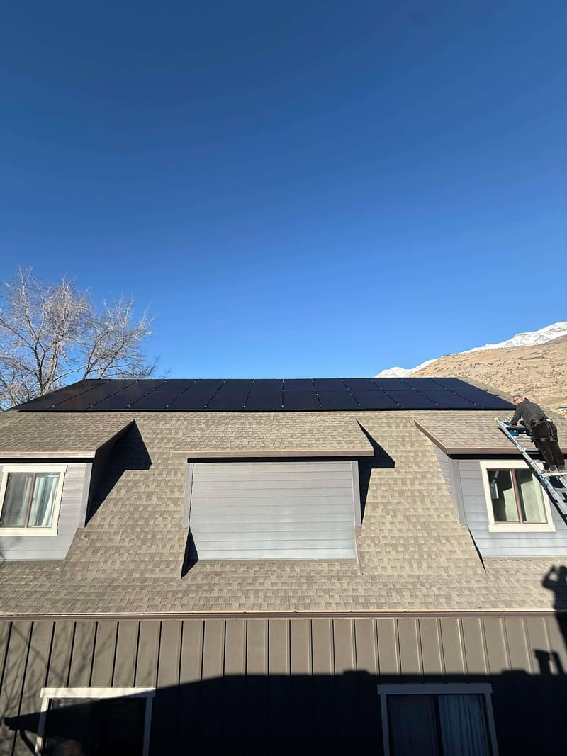 [Trina panels] Front view of house with full array of black solar panels on roof, installer on ladder, snow-capped mountains and blue sky behind