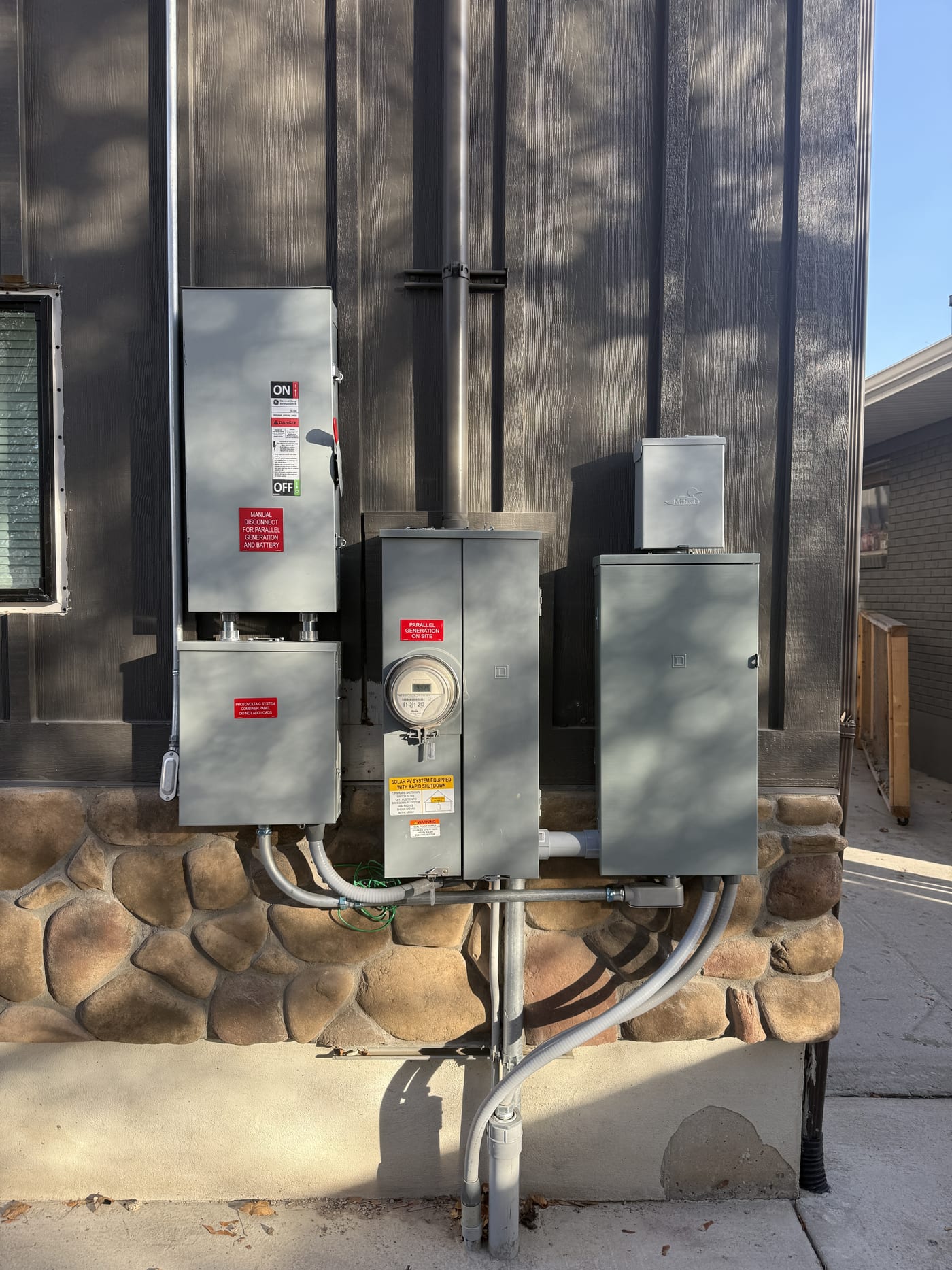 Slightly different angle of electrical equipment array with meter, panels, disconnects, and conduit on exterior wall