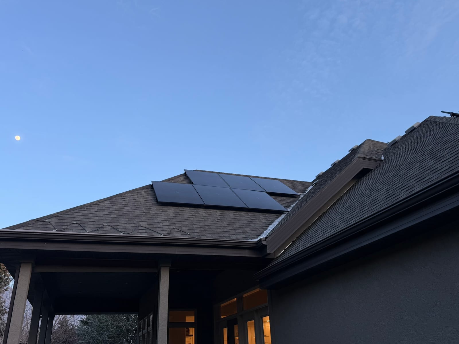 Stunning dusk shot of dark home with solar panels on roof, moon visible in deep blue sky, warm interior lights glowing through windows and porch, dramatic twilight atmosphere